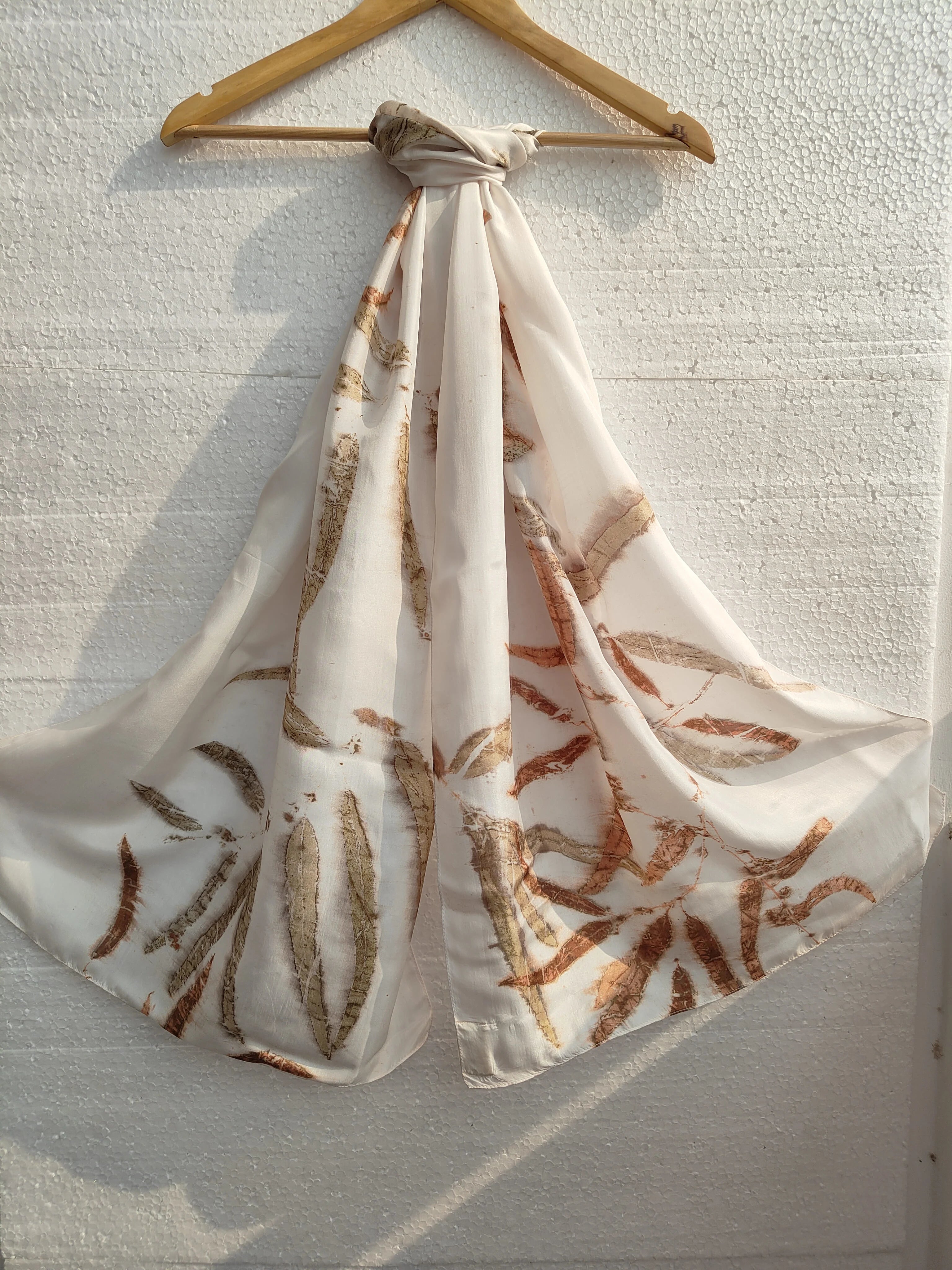 White silk scarf with brown Eucaplytus pattern draped over a wooden hanger against a textured white wall.
