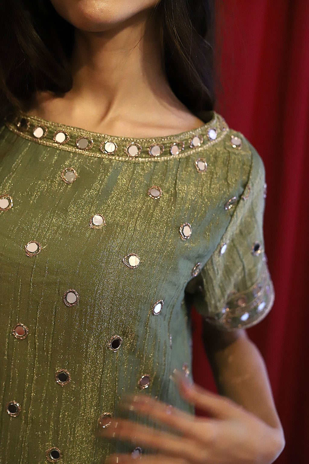 A close-up of a woman wearing a short-sleeved olive green dress decorated with silver mirror work mini dress, set against a dark red curtain background.