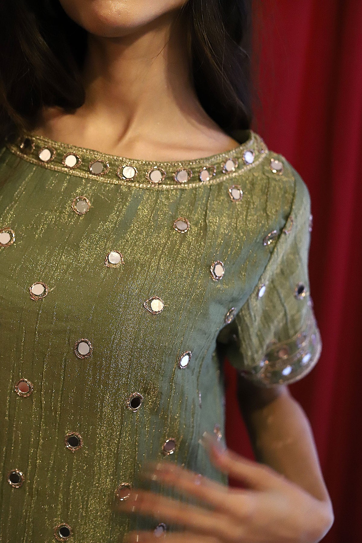 A close-up of a woman wearing a short-sleeved olive green dress decorated with silver mirror work mini dress, set against a dark red curtain background.