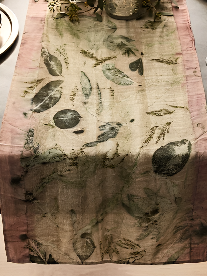 A rectangular eco printed table runner with a distressed botanical pattern, featuring muted green and black leaf motifs on a beige base with dusty pink borders, displayed on a dining table.