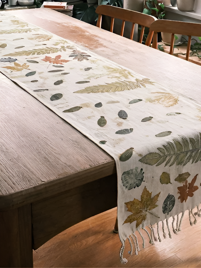 A cream table runner with a botanical leaf pattern in muted green, beige, and rust tones, draped over a wooden dining table with fringe at one end.