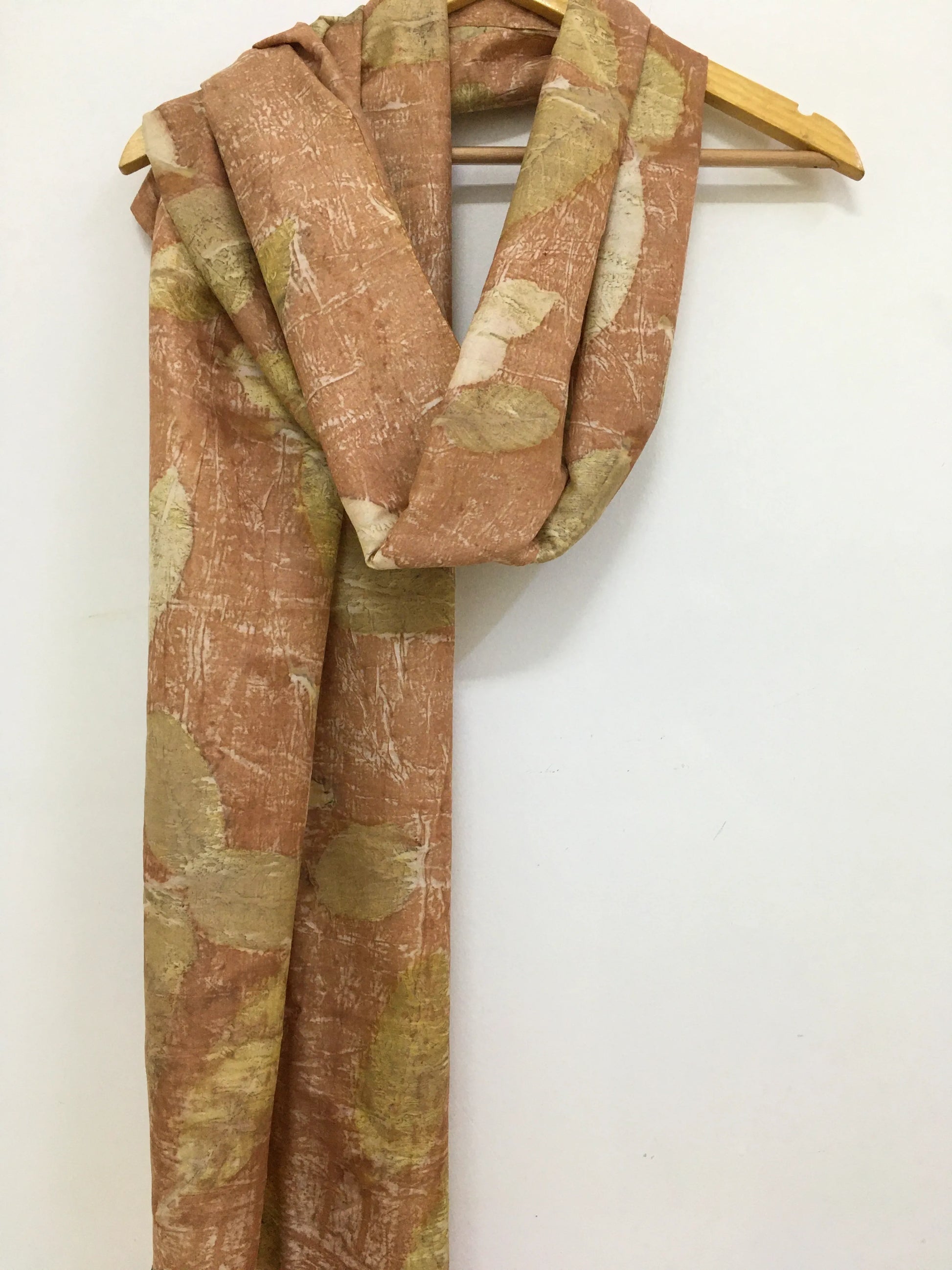 Brown and green patterned eco printed scarf on a wooden hanger against a white background