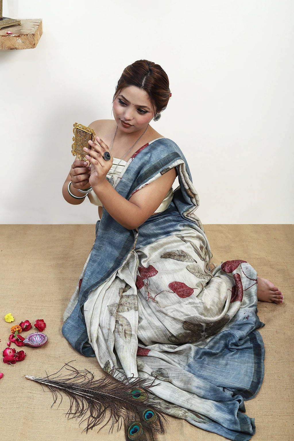 Woman in a traditional ecoprinted saree outfit sitting on the floor with a phone, surrounded by decorative items.