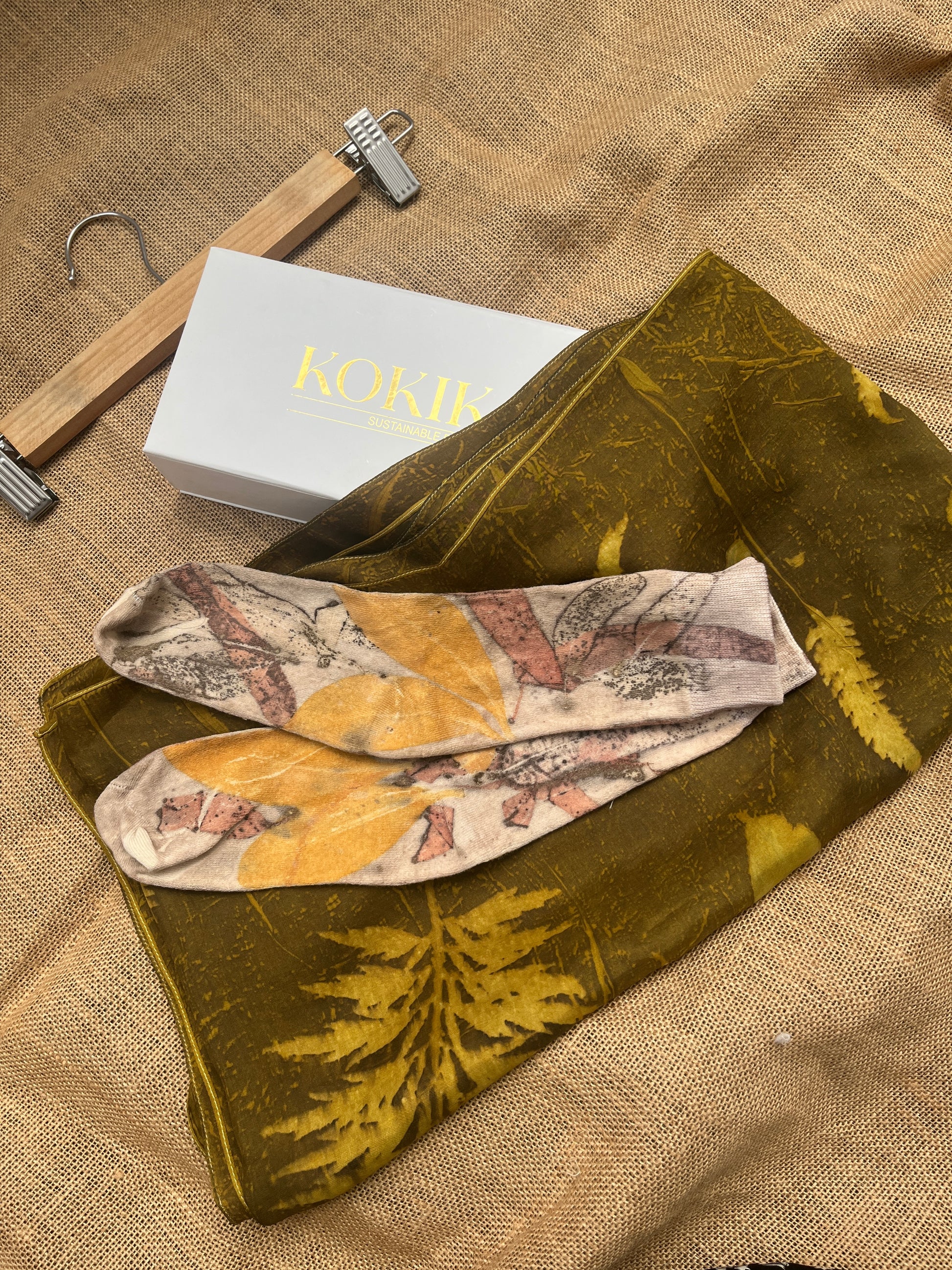 Folded floral-patterned socks on a green fabric with a KOKIKAR box and hanger in the background.