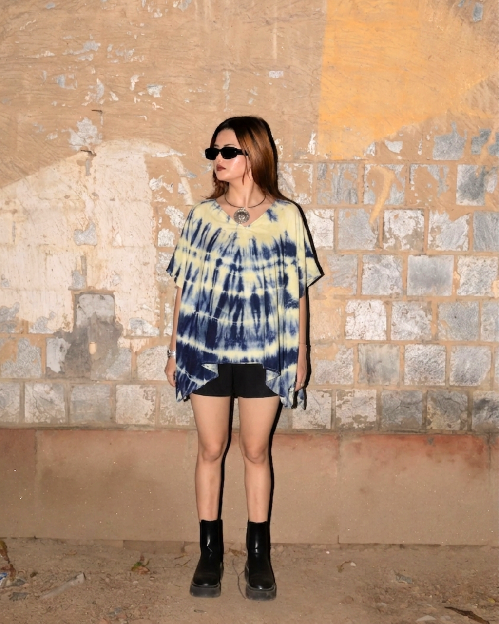 girl wearing a tie dye Kaftan Flared Top and black shorts standing against a textured wall.