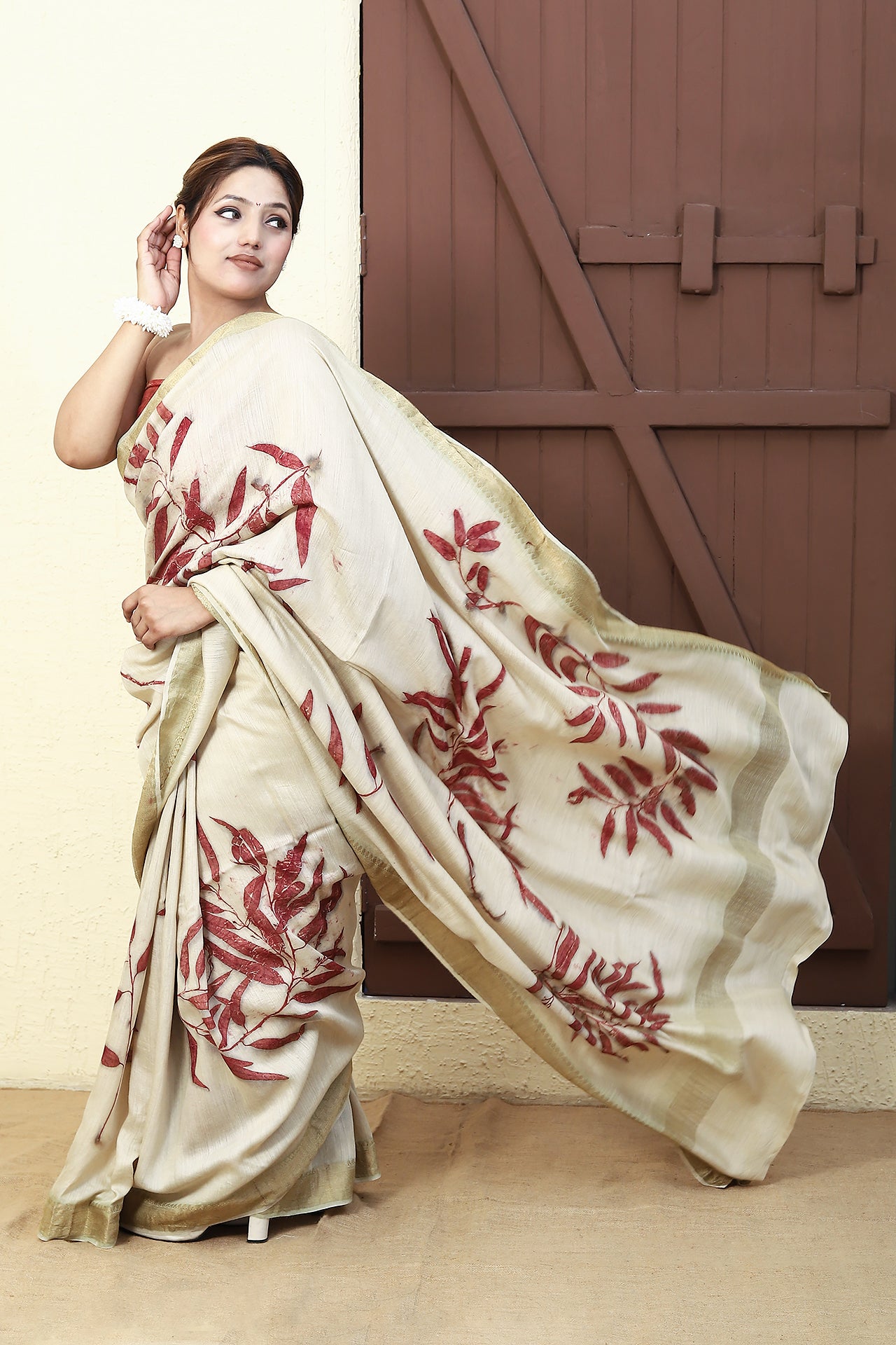 Woman wearing a white Madder Drape Muga Silk Saree with red floral patterns in front of a wooden door.