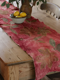 A rustic ecoprinted table runner with a distressed leaf pattern draped over a wooden dining table, styled with a small bowl of lemons and a vase in the background.