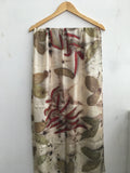 A long scarf with a beige base, featuring a botanical pattern in muted green and brown, overlaid with large red madder eucalyptus leaves, hanging on a wooden hanger against a white wall.