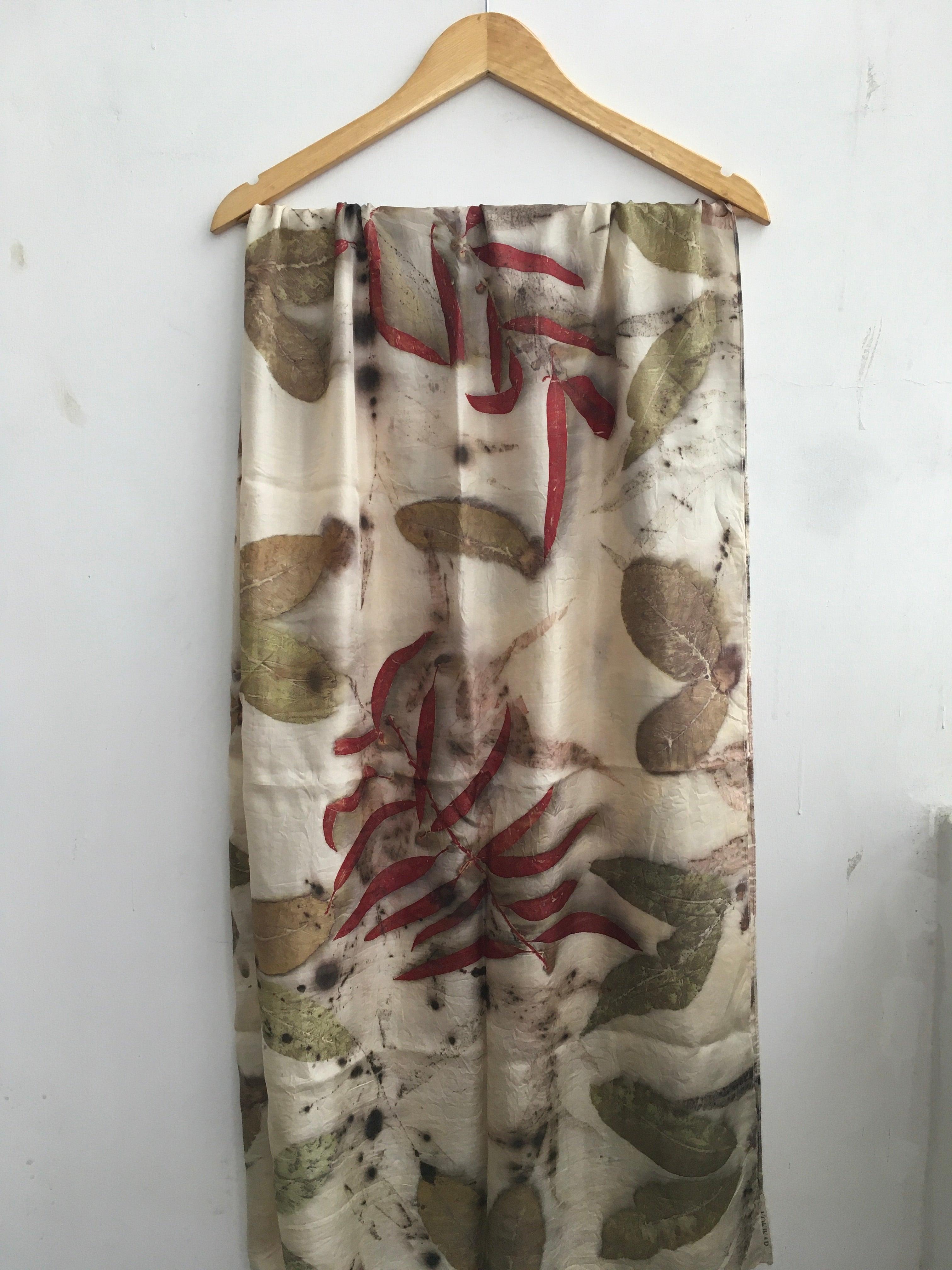 A long scarf with a beige base, featuring a botanical pattern in muted green and brown, overlaid with large red madder eucalyptus leaves, hanging on a wooden hanger against a white wall.