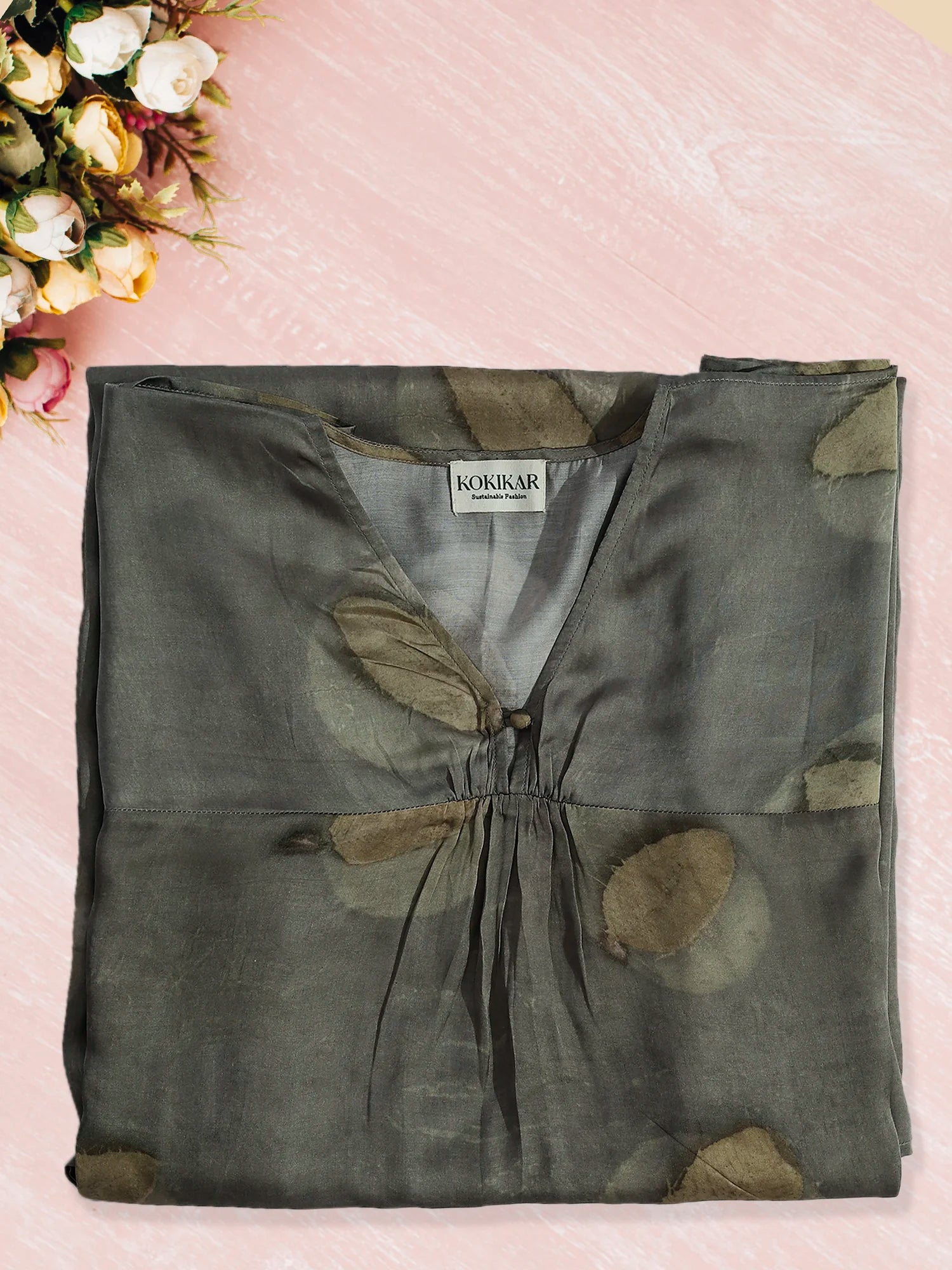 Folded green ecoprinted kaftan with a kokikar brand label on a pink surface