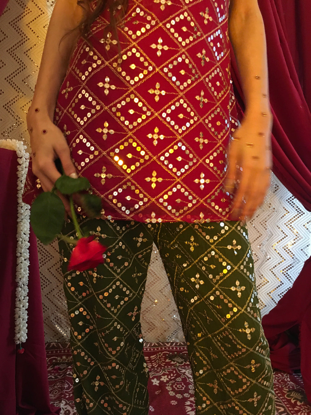 A close-up of a person wearing traditional embellished fabric garments, with one leg in deep red and gold patterned fabric and the other in dark green with gold embroidery. The person is holding a red rose, with draped red fabric and patterned textiles in the background.