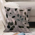 A square decorative ecoprinted  cushion cover with a gray and lavender watercolor background and dark green leaf patterns, styled on a sofa next to a white textured blanket.