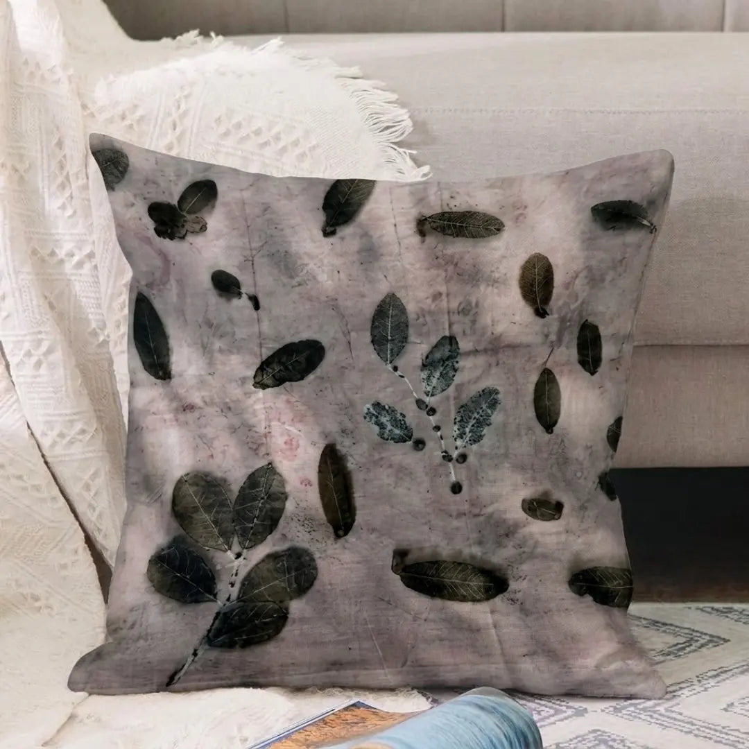 A square decorative ecoprinted  cushion cover with a gray and lavender watercolor background and dark green leaf patterns, styled on a sofa next to a white textured blanket.