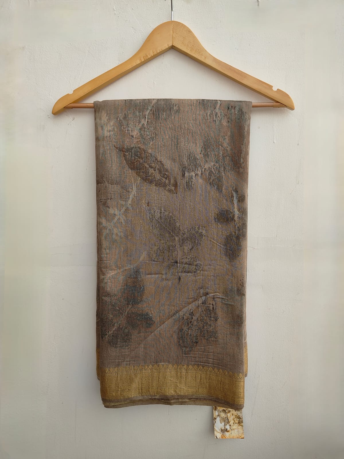 A Muga silk saree with leaf patterns, displayed on a wooden hanger, with a beige and brown color scheme.