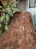 Dark brown pure silk scarf naturally dyed with catechu and eco printed with seasonal botanical leaves using sustainable techniques draped over a chair next to a green plant.