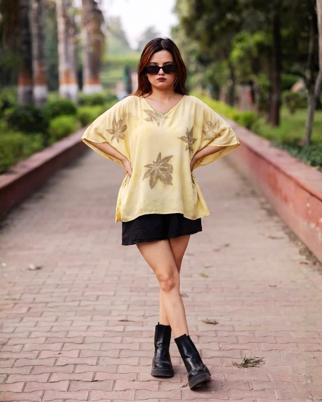 Woman in a yellow Kaftan Flared Top and black skirt standing on a brick path with greenery in the background