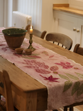 A rustic dining table in a warm kitchen setting, decorated with a pink linen ecoprinted table runner featuring a leaf and flower pattern, alongside a green ceramic bowl and a lit candle.