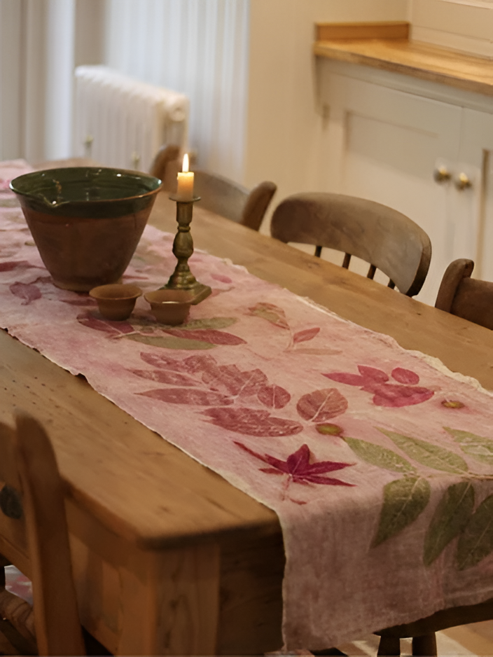 A rustic dining table in a warm kitchen setting, decorated with a pink linen ecoprinted table runner featuring a leaf and flower pattern, alongside a green ceramic bowl and a lit candle.