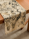 An off-white ecoprinted table runner with a dark gray and black abstract leaf-like pattern and fringed edges, draped over a wooden table.