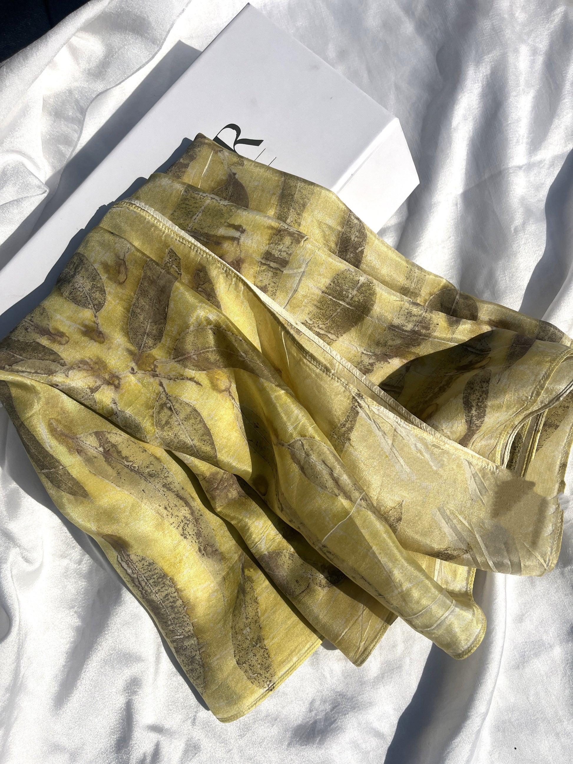 A yellow silk scarf with brown eucalyptus leaf prints, displayed on a white surface inside a box.