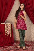 A woman models a traditional South Asian outfit consisting of a sleeveless magenta kurta with white geometric patterns and dark green flared pants with white motifs, set against a backdrop of red curtains and a patterned rug.